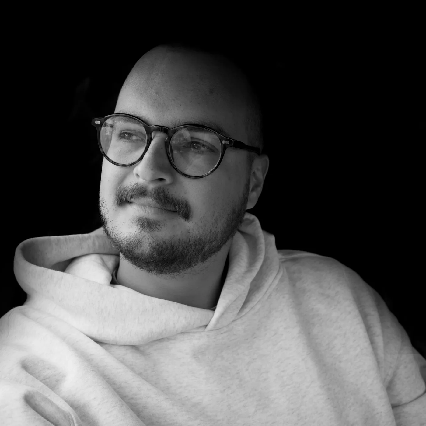 Black and white portrait of a person with a shaved head, glasses, and a moustache and beard, wearing a light hoodie and looking off to the side against a dark background.