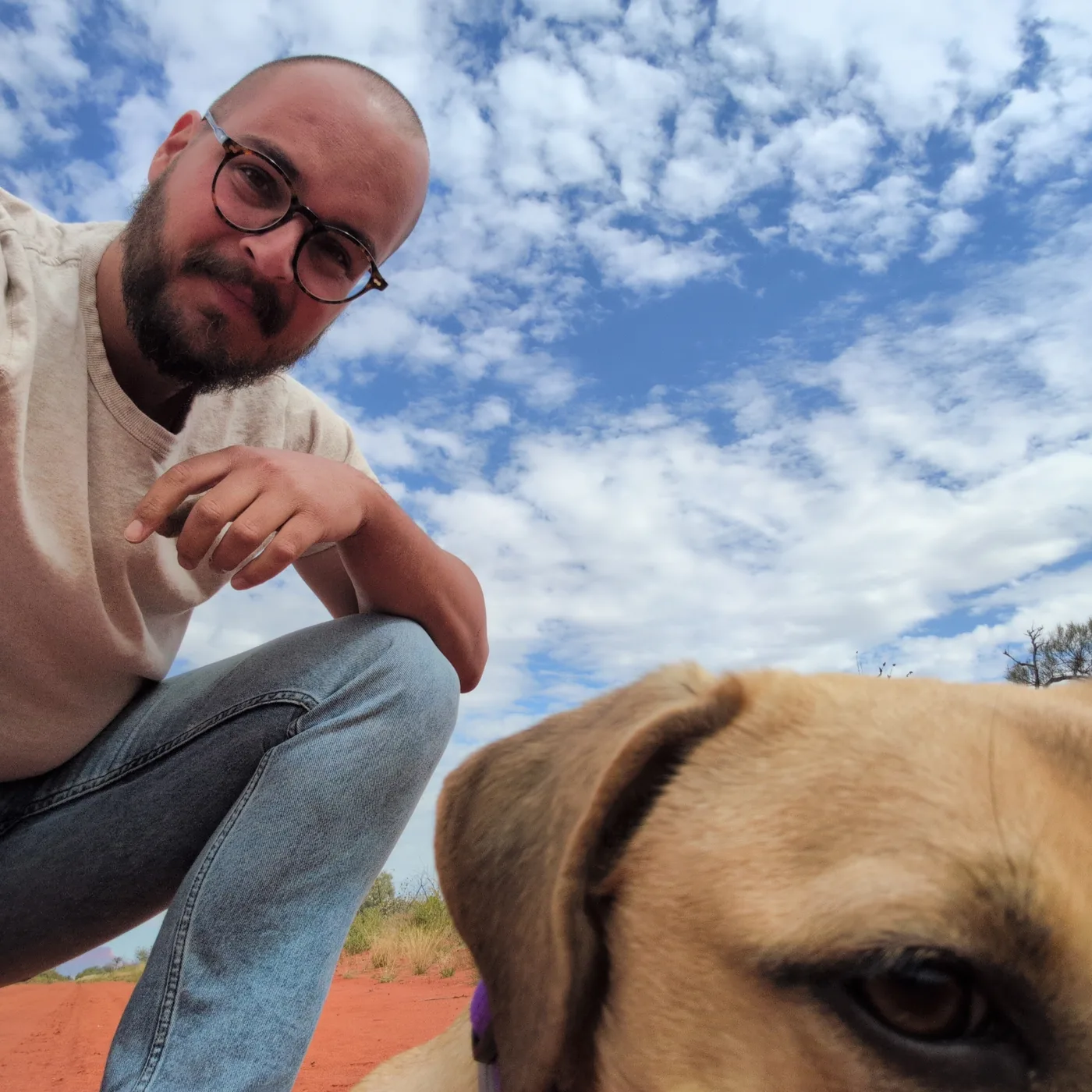 A person with a shaved head, dark beard and round glasses crouches on red dirt under a blue sky full of scattered cloud, looking down at the camera. A tan dog's face fills the bottom right corner of the frame, cropped at the eyes.